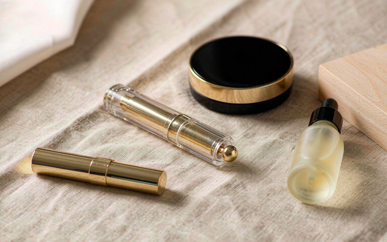Assortment of cosmetic packaging components — lipstick case, gloss tube, compact and glass bottle — laid out on a light surface for comparison